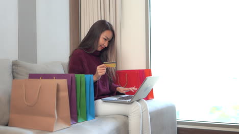 asian women surrounded with shopping bags using her credit card for shopping online, type in card information on the laptop, showing the winning expression