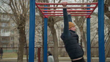 A coach jumps up to grab a red iron bar in an outdoor workout area, with people and building with bare trees in the background