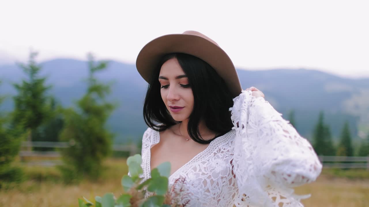 Attractive bride in hat on mountainous background. Beautiful woman in white dress with bouquet of flowers standing among nature and looking aside.