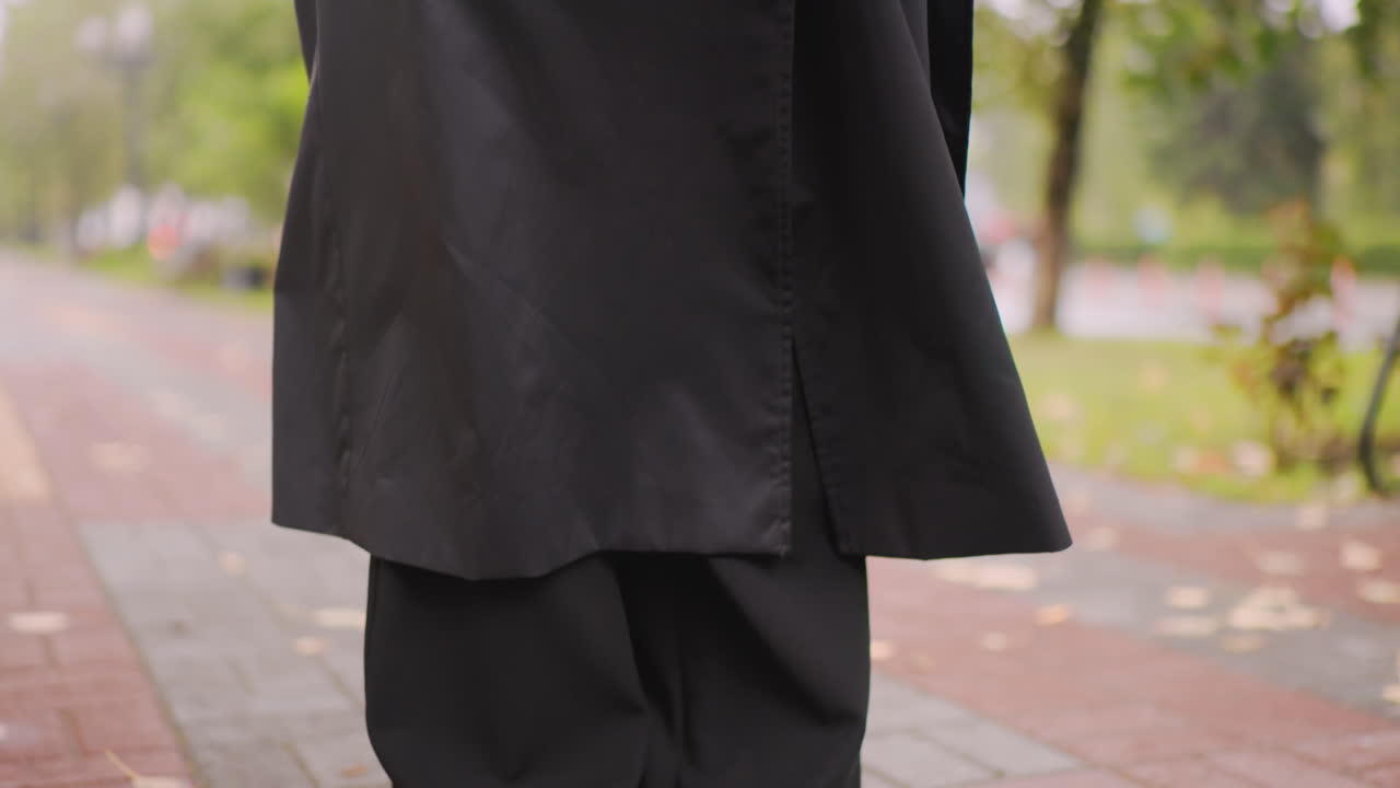 Close-up of black trench coat with large buttons worn outdoors on rainy autumn day, blurred green park and pavement in background, detail of stylish outerwear showing fabric texture and seasonal fashion