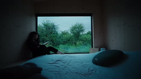 woman reading a book during dusk by window in a cabin in nature wide shot