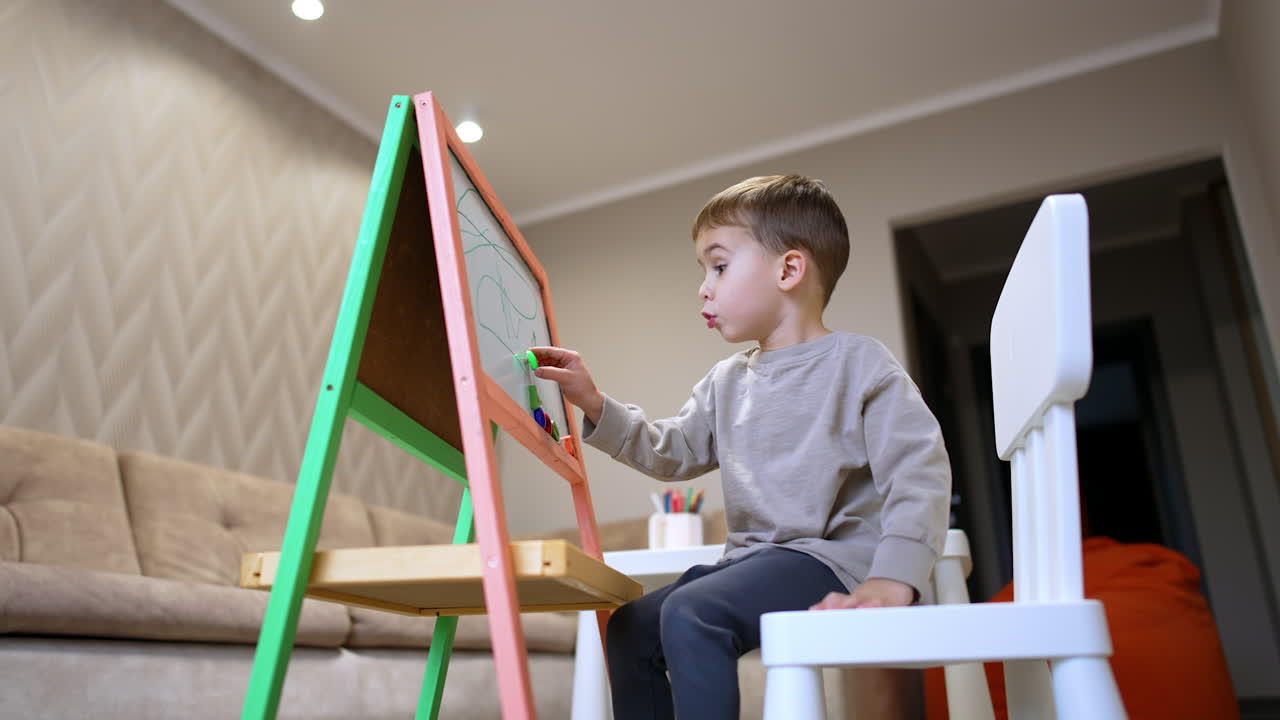 Young child focused on drawing at easel. A young boy sits on a chair, engrossed in drawing on a colorful easel in a cozy living room setting