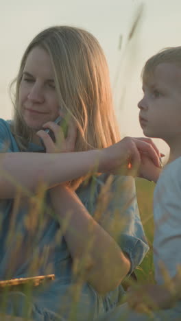 a mother in a blue gown multitasks in a grassy field, using a laptop and phone, with her two young sons beside her, one in yellow engrossed in the laptop,the other in white,playfully touching her hair