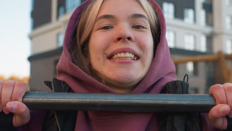 portrait of cheerful young woman in hoodie winking and smiling while pulling up on horizontal bar during outdoor fitness in urban park, showcasing energetic strength and calisthenics workout