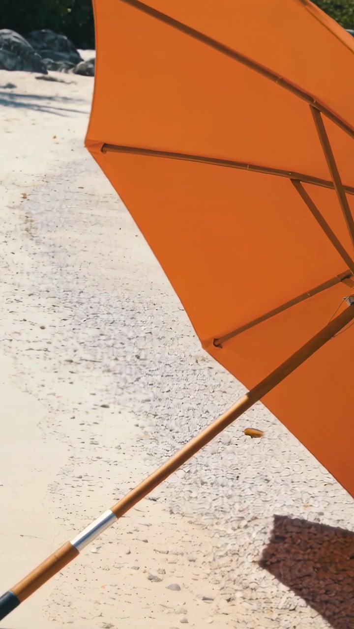 Video verticale: Telecamera panoramica che mostra un ombrellone da spiaggia arancione ancorato nella sabbia, rivelandone la trama.
