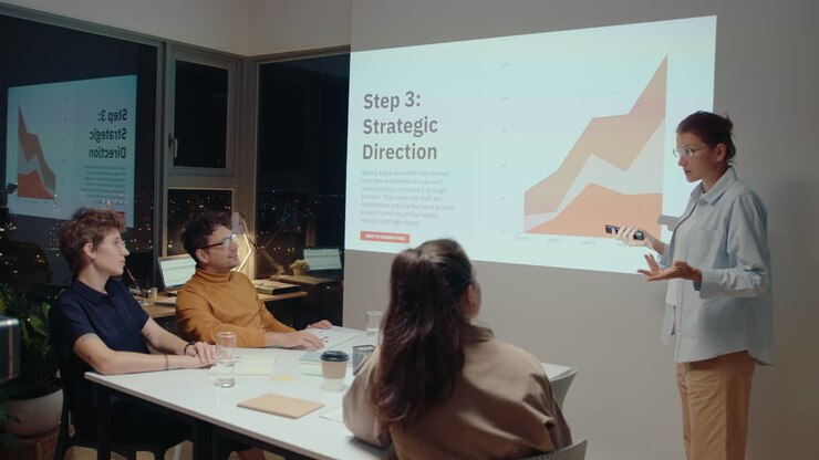 mujer de negocios de pie junto a la pantalla de proyección frente al equipo, cambiando diapositivas con control remoto, dando una presentación durante una reunión en una oficina con ventanas panorámicas con vistas al paisaje urbano nocturno