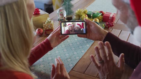 Pareja-Caucásica-Con-Gorros-De-Papá-Noel-Usando-Un-Teléfono-Inteligente-Para-Una-Videollamada-Navideña-Con-Una-Pareja-En-La-Pantalla