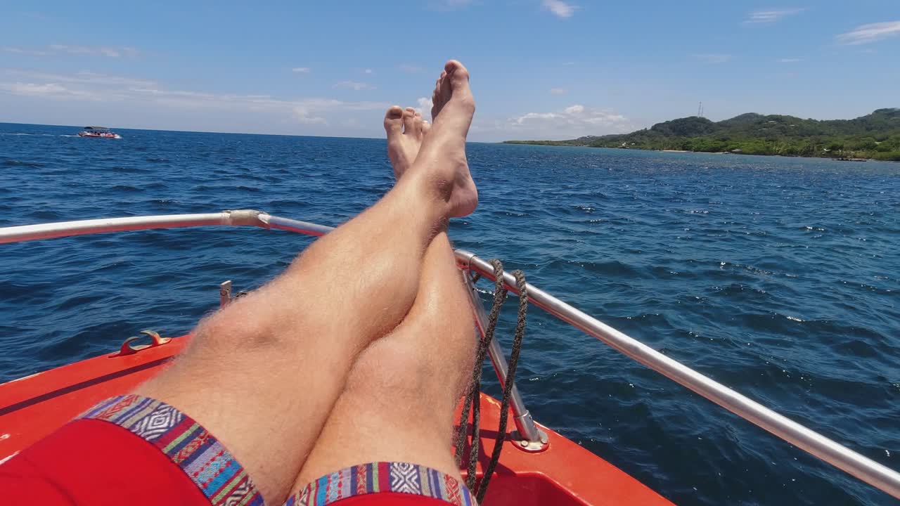 pov: piernas masculinas peludas en la proa de un barco rojo en aguas azules oscuras cerca de la costa