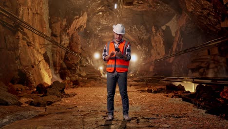 full body of asian male engineer with safety helmet using smartphone while standing in underground mine tunnel