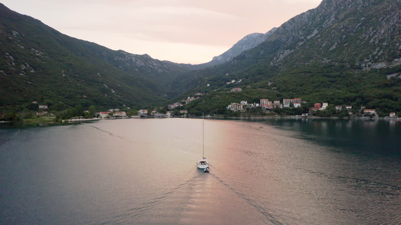 Uno yacht bianco lussuoso con un alto albero senza vele che traina un motoscafo vuoto e si dirige verso il porto di una piccola città costiera nella baia di Kotor in Montenegro, con un bagliore rosso sull'acqua e sulle montagne circostanti