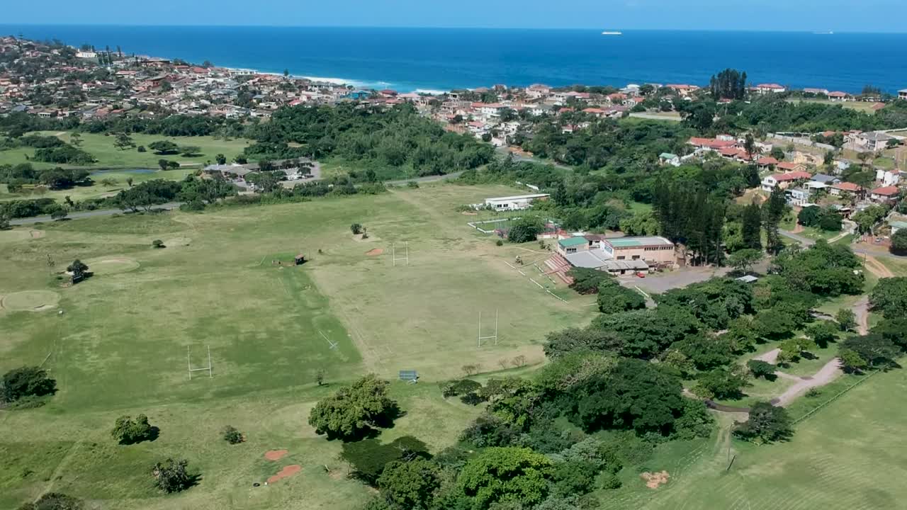 Drone in ascesa con una chiara vista di un campo sportivo di rugby e calcio circondato da case residenziali e una vista del mare all'orizzonte
