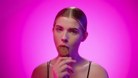 young adult blonde caucasian girl eats an ice cream stick in a pink studio with chroma background