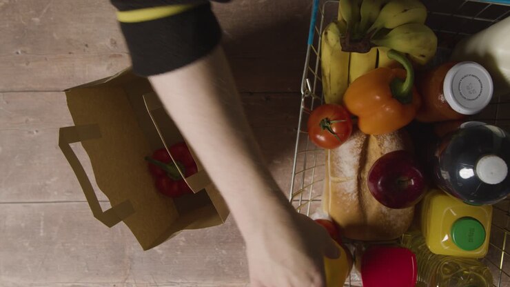 Overhead Shot Of Person Packing Basic Fresh Food Items From Supermarket Wire Shopping Basket Into Paper Bag
