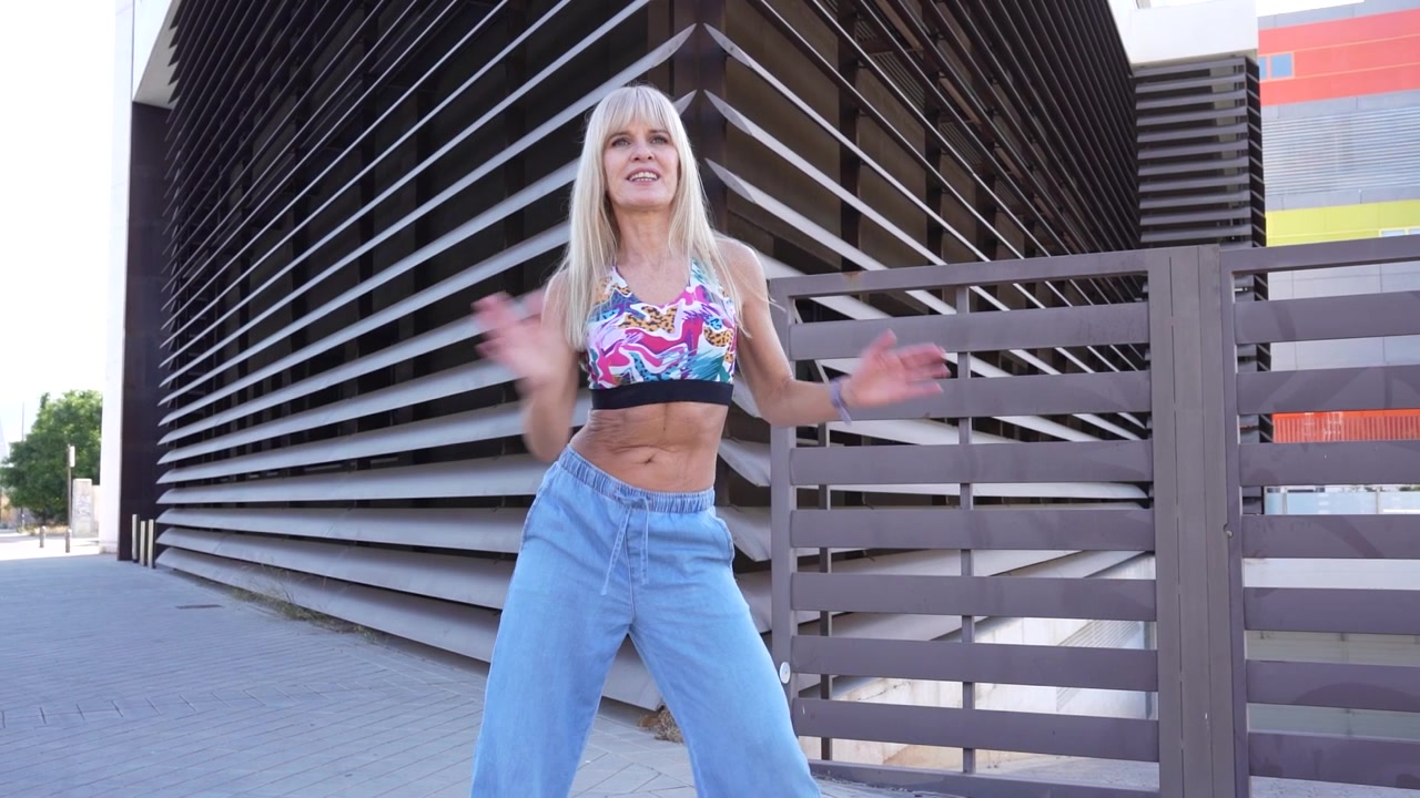 Blonde woman in colorful top and jeans poses outdoors beside modern architecture