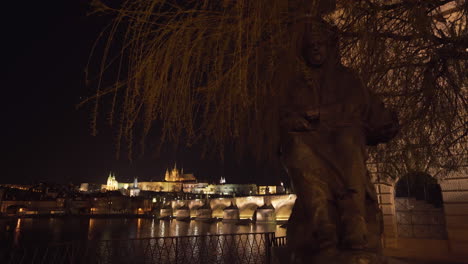 El-Castillo-De-Praga-Y-El-Puente-Charles-Sobre-El-Río-Vltava-En-El-Centro-Histórico-De-Praga,-Chequia,-Iluminados-Por-Luces-En-La-Noche,-Disparados-Desde-Debajo-De-Una-Estatua-De-Un-Rey-Y-Un-árbol-Al-Otro-Lado-Del-Río