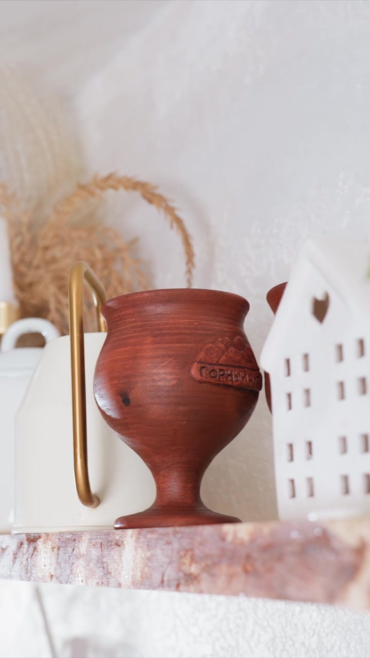 Estante con tazas de madera, una taza de cerámica blanca con forma de casa. Objeto decorativo de casa blanca con un recorte en forma de corazón. El entorno incluye una pared blanca texturizada y varios elementos decorativos.