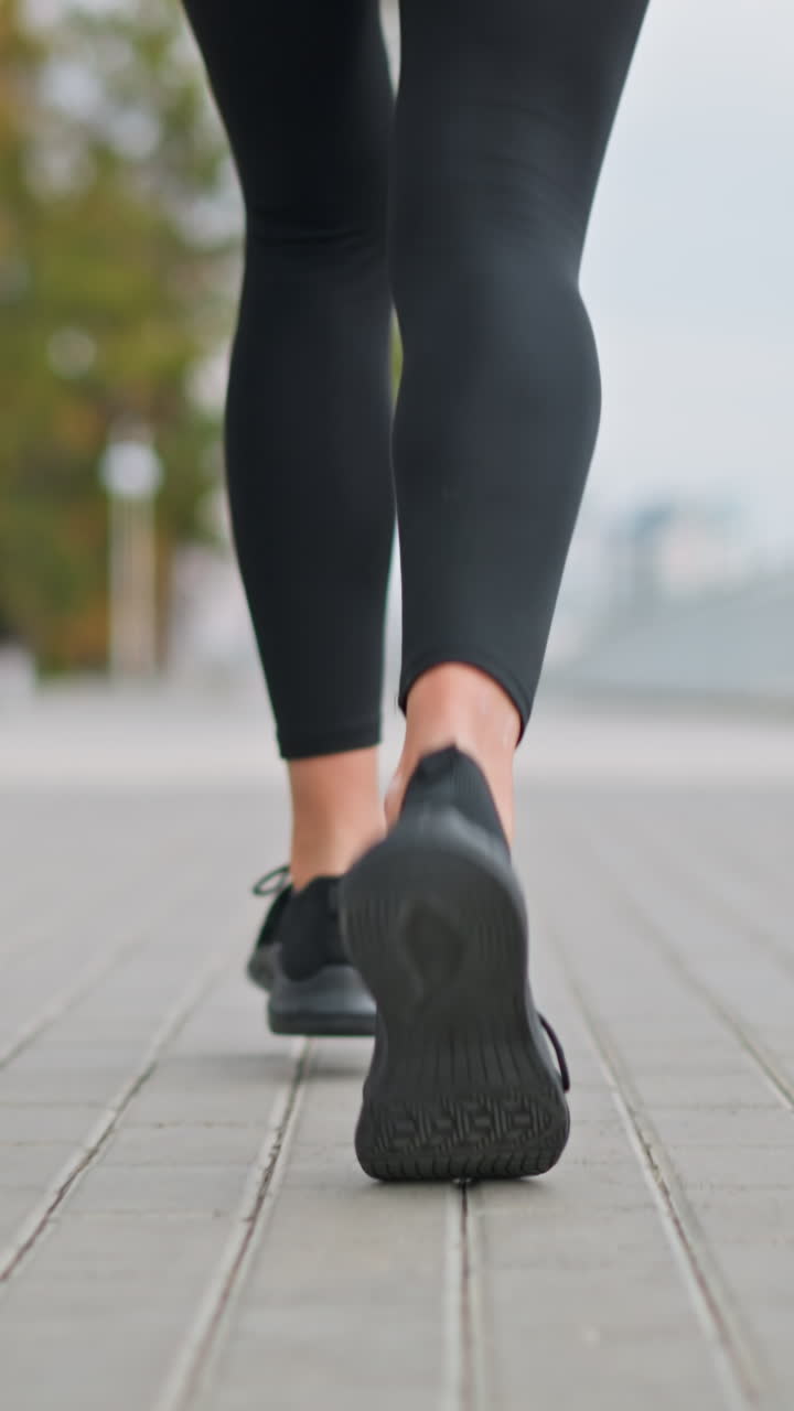 Close-up view of woman jogging in sneakers along paved street path near iron fence with blurred background showing trees and distant landscape under cloudy sky