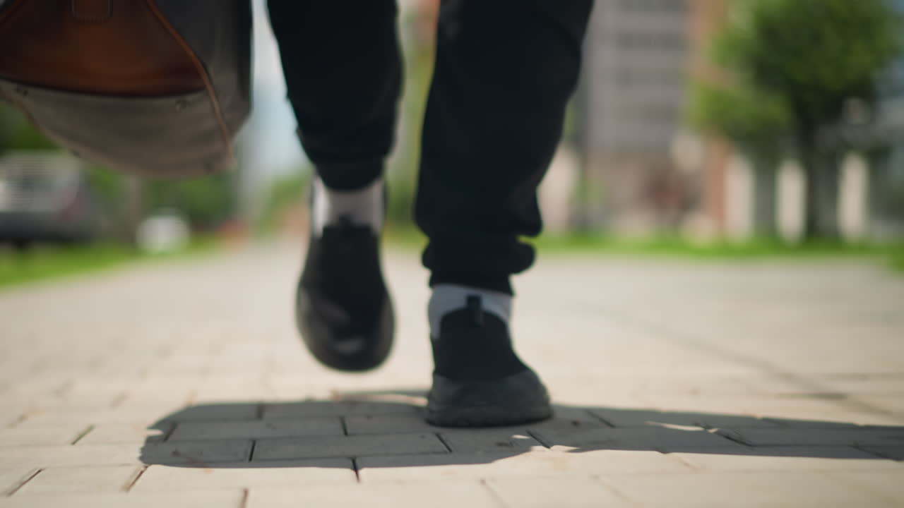 도시 학생의 통학, 도시 환경 속 캐주얼 스니커즈, 캠퍼스 길을 따라 자신감 있게 걷는 젊은이, 도시 환경에서 녹색 경계를 지나 운동화를 신고 이동하는 사람