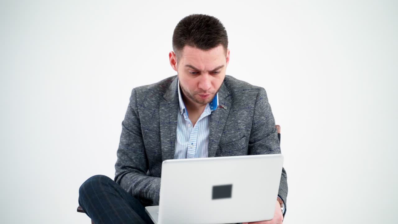 Grieved businessman looking into the laptop. Pensive male freelancer working on a modern gadget while sitting alone on white background.