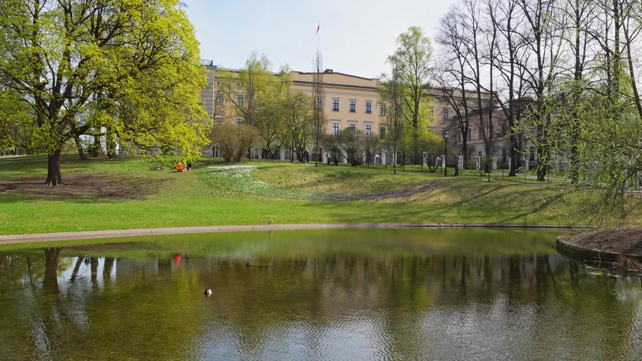 Plano general de un lago tranquilo en el centro de Oslo con un paisaje diverso de formaciones esponjosas. Reflejos de un edificio amarillo histórico, vegetación fresca y árboles primaverales se bañan en la suave luz del día, sin personas visibles.
