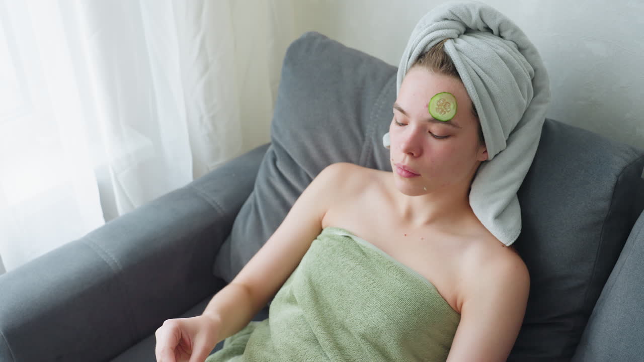 Woman relaxing on couch, wrapped in towel, placing cucumber slice on her face to refresh skin in a calm, bright atmosphere with soft sunlight streaming through a window