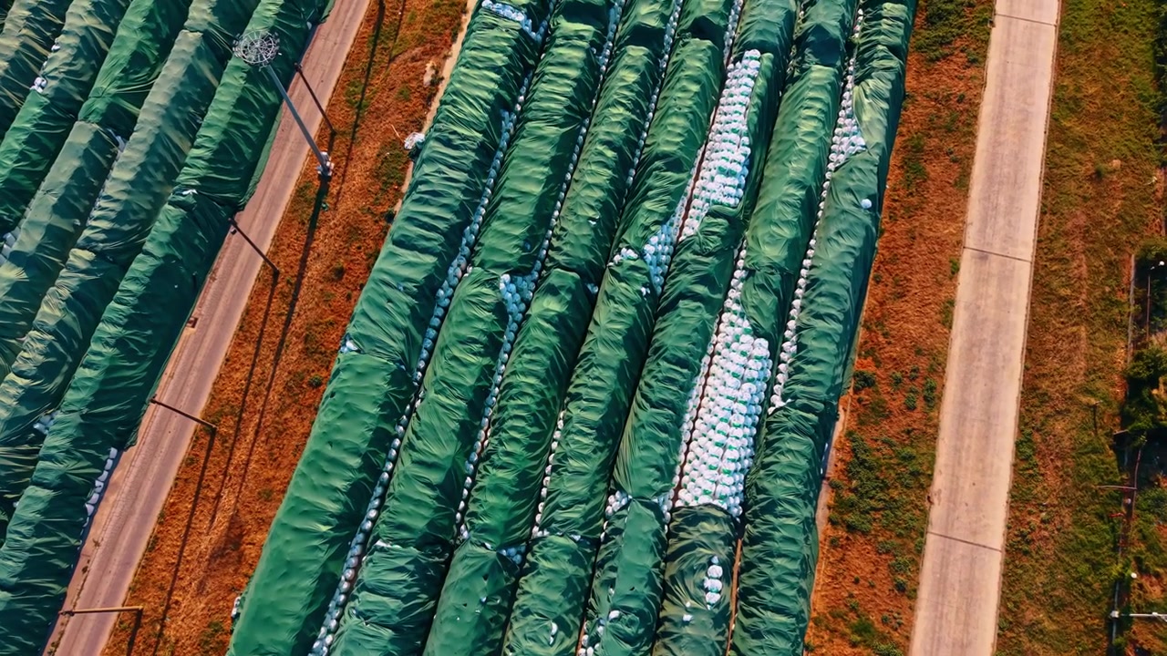 Piazzale di stoccaggio agricolo, pile di sacchi coperti con teli verdi. Prospettiva dall'alto che cattura file parallele di materiali stoccati separati da percorsi in cemento durante il giorno.