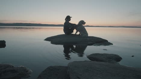 woman petting dog on rock in lake at sunset