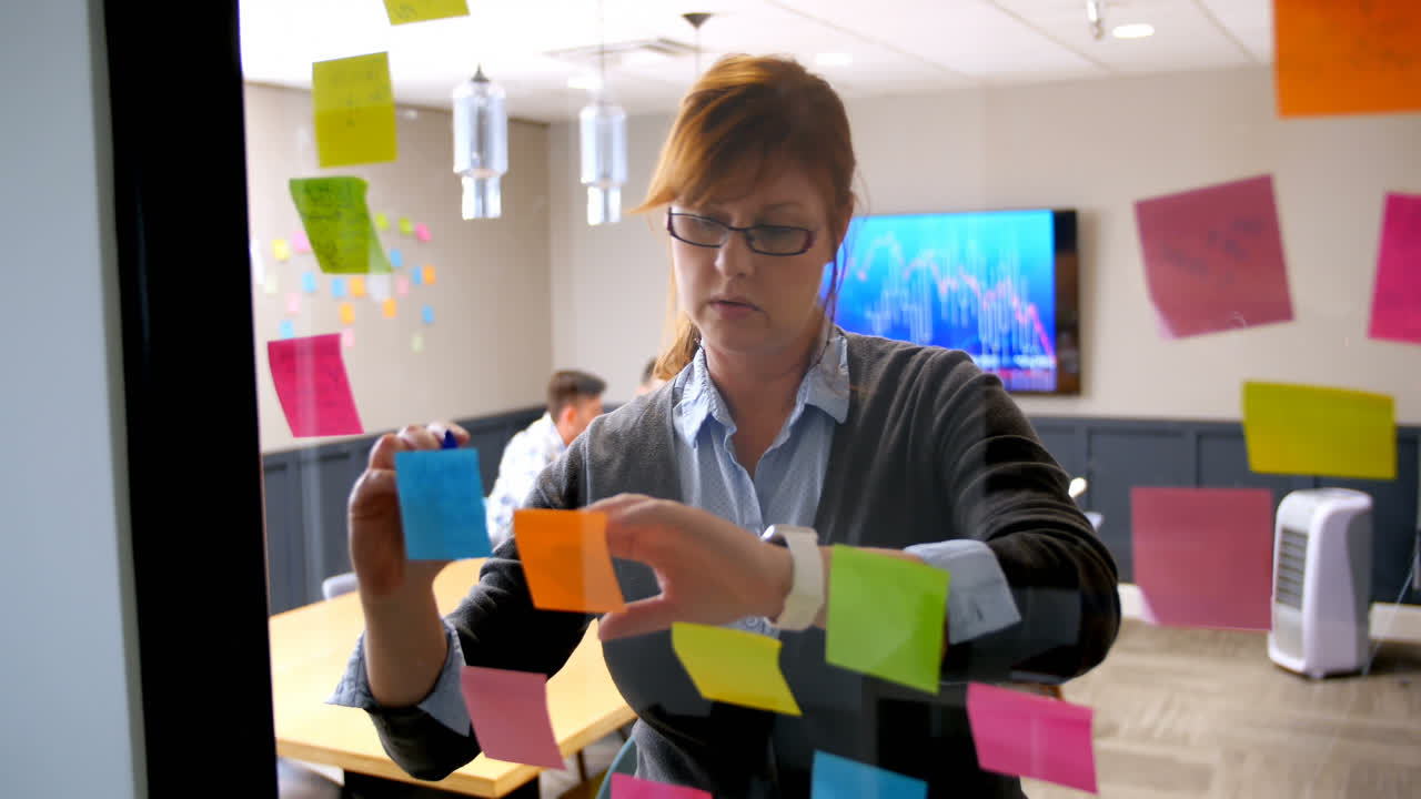Female executive using smartwatch while writing on sticky note on glass wall 4k