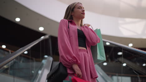 lady wearing a black crop top under a pink outfit, holding a mint green shopping bag behind her and another bag in her right hand while in an escalator moving upward, in a shopping mall interior