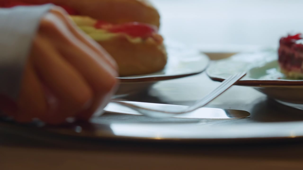 Close up of hand reaching for silverware on plates with food in background. focus shifts between forks and knives as they are picked up revealing blurred details of sandwiches or desserts