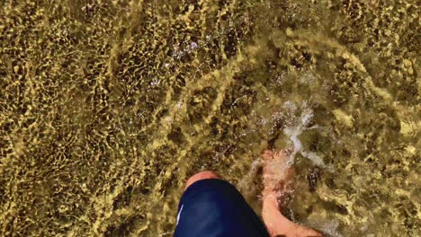 caucasian male legs and feet walking through ocean waves barefoot towards sandy beach