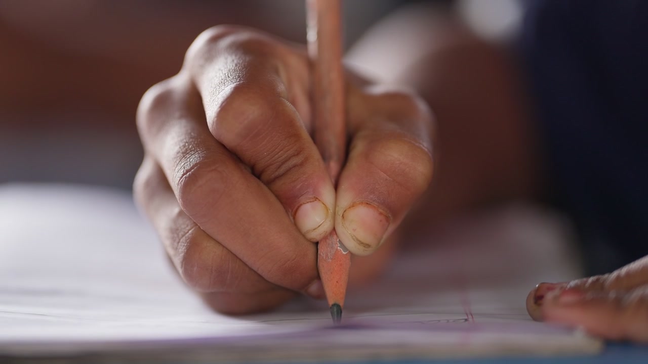 Close-up of a student's hand gripping a pencil while writing on white paper in rural India. Shallow depth of field isolates the writing hand against a soft-focused background.
