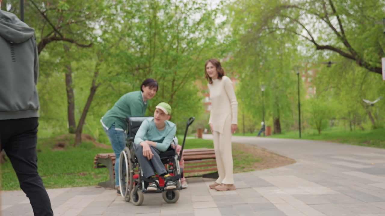 Park Gathering Around Wheelchair While Stroller Passes By, Adults Assisting And Chatting, Toddler Near Feet, Leafy Path, Stroller Silhouette In Foreground, Inclusive Urban Park Moment, Supportive