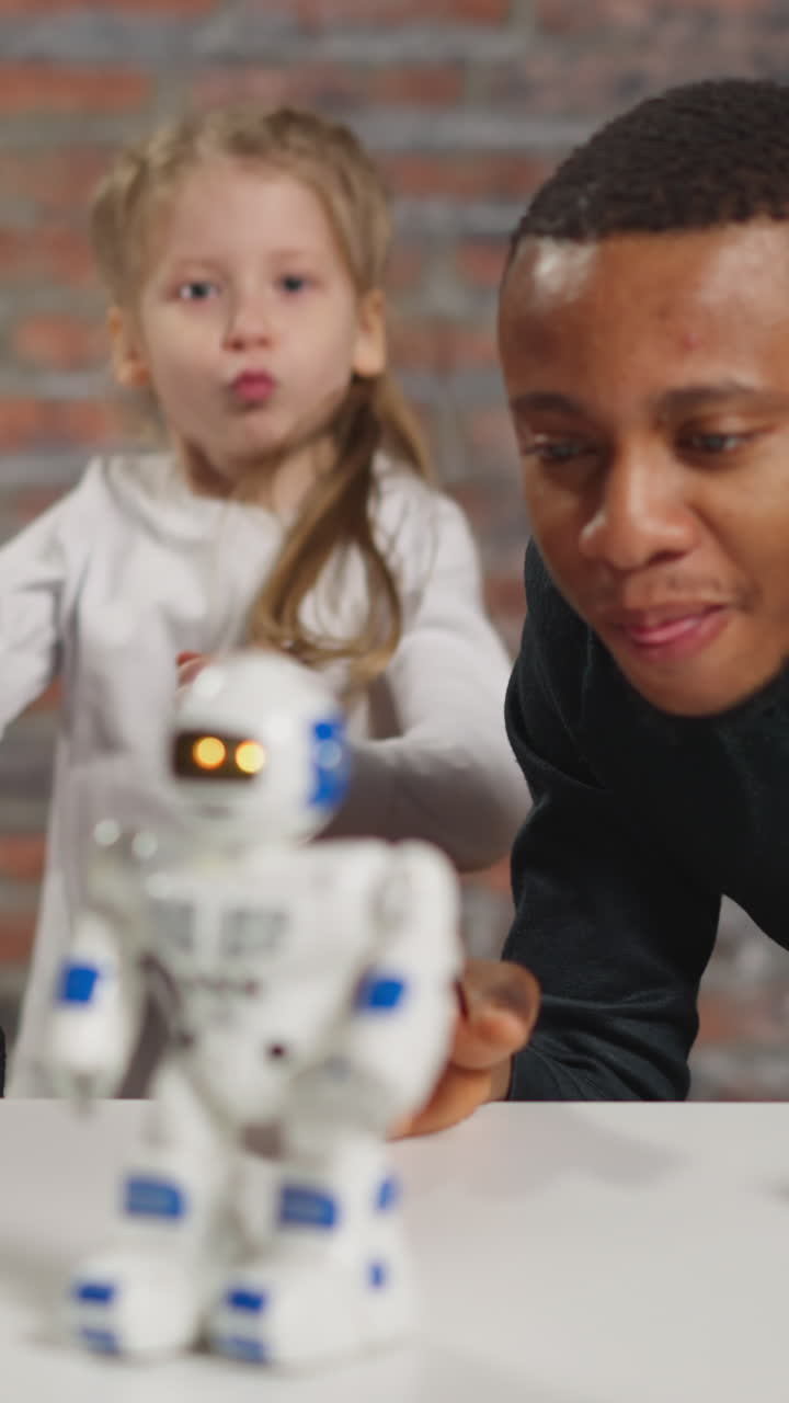 Interested man with positive Asian colleague look at moving robot on table while schoolgirl dances in spacious studio