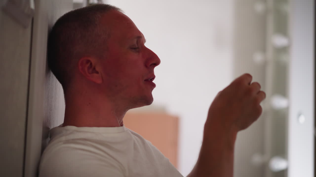 side view of middle aged man seated against wall wearing white shirt enjoying eating chicken savoring taste in relaxed indoor setting with soft lighting and blurred background reflection