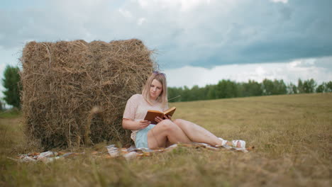 lady sits on blanket in open farmland, reading brown-covered notebook with hay bale behind, vast countryside landscape with cloudy sky and green trees in distance