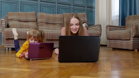 Mother-and-small-daughter-child-sit-on-warm-wooden-floor-at-home-working-on-laptop,-watching-cartoon