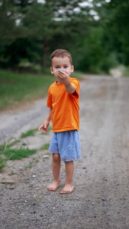 little barefoot toddler boy walking by the ground road stops and turns around. kid picks up a stone, shows it to the camera and throws to the side. vertical video.