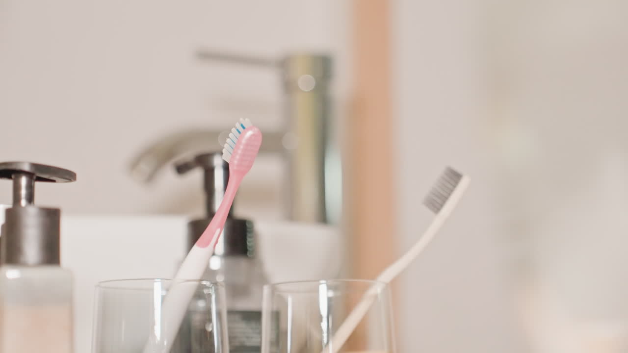 Primer plano de los cepillos de dientes en el lavabo del baño del hotel. El personal de limpieza coloca un cepillo de dientes para el huésped; unas manos eficientes disponen un cepillo rosa en un vaso cerca del dispensador de jabón. Se observa un rápido movimiento de limpieza para la preparación de la habitación.