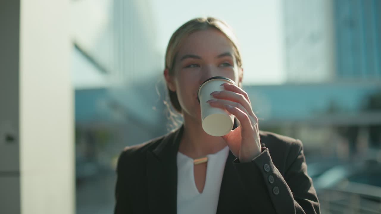 Woman with refreshing look in professional attire walks by modern glass building holding tablet in one hand and takeaway tea in other hand, sipping drink calmly in bright urban outdoor setting