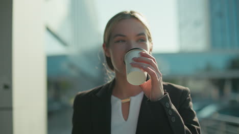 woman with refreshing look in professional attire walks by modern glass building holding tablet in one hand and takeaway tea in other hand, sipping drink calmly in bright urban outdoor setting