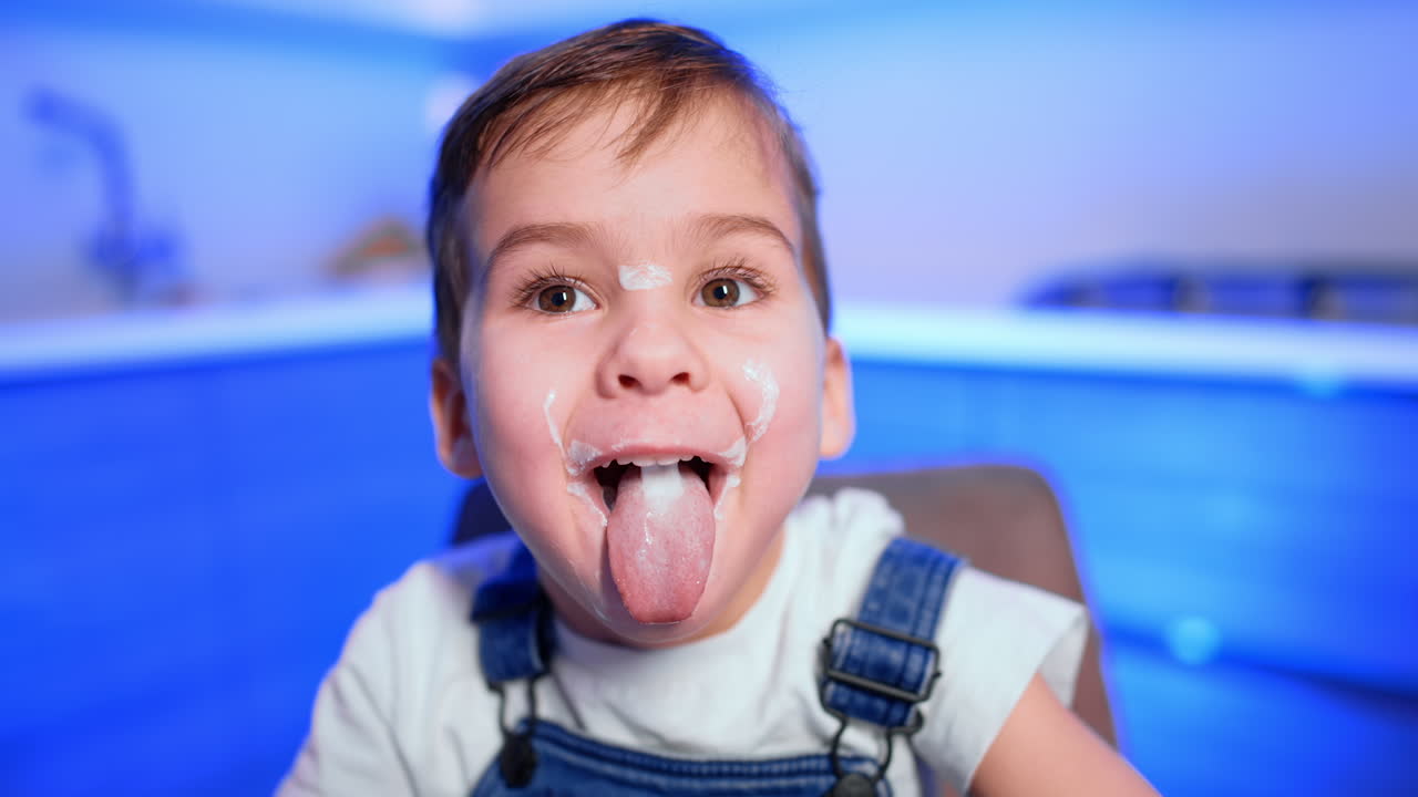Caucasian baby boy with smudged face sits at kitchen table. Funny kid has eaten yogurt and shows his tongue. Close up.