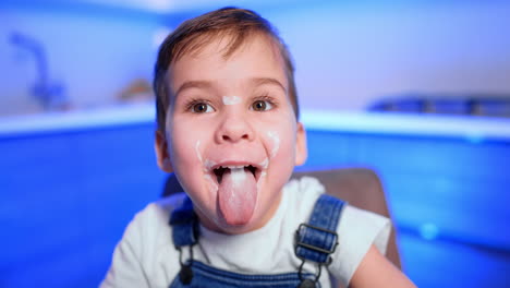 caucasian baby boy with smudged face sits at kitchen table. funny kid has eaten yogurt and shows his tongue. close up.