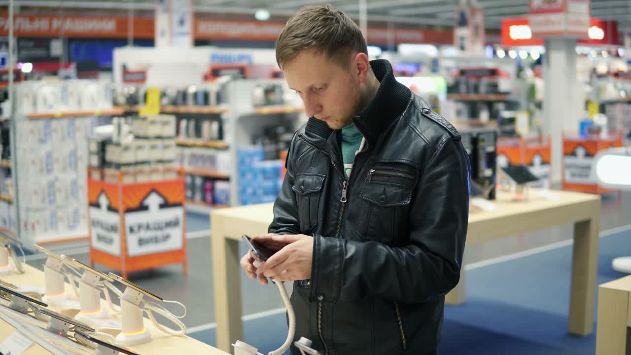 Young man in black leather jacket is choosing a new mobile phone in a shop, checking how it works