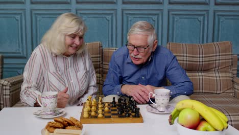 smiling senior couple grandfather grandmother resting on sofa drinking coffee, playing chess at home
