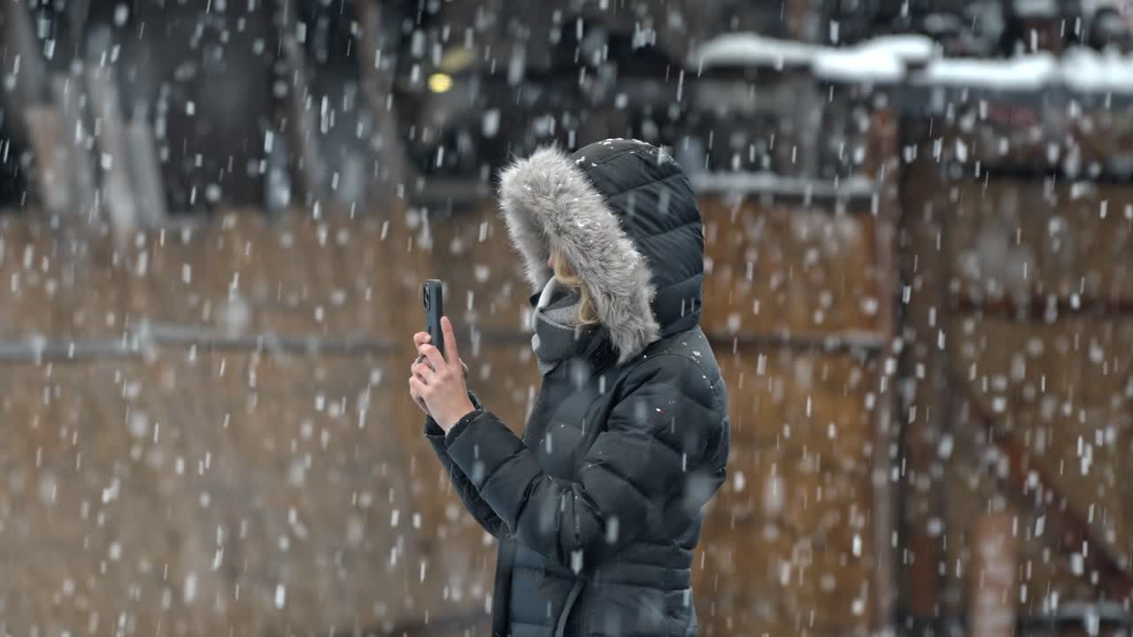 A woman captures the beauty of a snow-covered Shirakawa-go with her phone, as snowflakes gently fall around her.