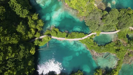 Take-off-over-the-Plitvice-Lakes-National-Park-with-many-green-plants-and-beautiful-lakes,-a-mountainous-road-along-which-people-walk