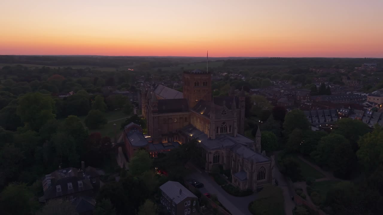 Aerial drone orbit around St Albans Cathedral at sunset, capturing the illuminated landmark with the city skyline glowing in the background under warm evening light