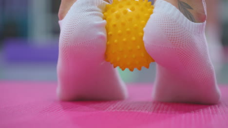 close-up of a person leg in socks slightly raised while holding a yellow spiky ball on a pink mat, performing a stamina build-up exercise with tattoo visible