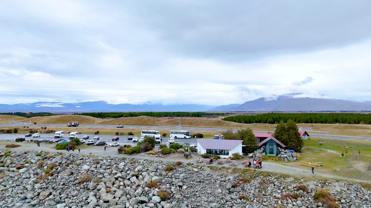 Aerial footage captures a serene landscape with scattered buildings and vehicles near Lake Pukaki under a cloudy sky